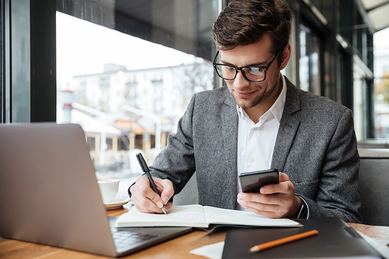 Файл:Smiling-businessman-eyeglasses-sitting-by-table-cafe-with-laptop-computer-while-using-smartphone-writing-something 171337-5589.jpg